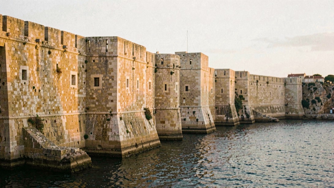 Sur les remparts nord d'Alghero au lever du jour, les bastions de pierre blonde éclairés de biais, avec les tours du golfe en arrière-plan sur une mer calme, cadrage large au 35 mm.