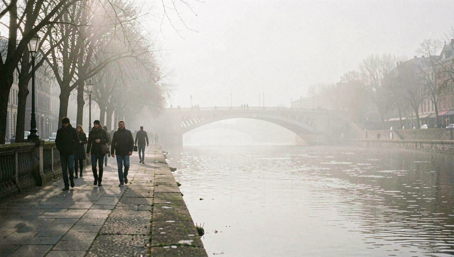 Quai le long d'un fleuve dans une brume légère, silhouettes de passants et arche de pont qui pâlit au fond