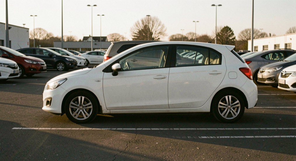 Berline compacte blanche à l'arrêt sur un parking de location au lever du jour, flanc gauche entier dans la lumière rasante, ombres longues sur le bitume, cadrage de profil à quelques mètres